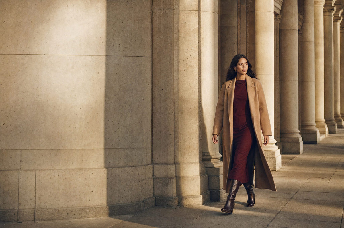 Woman in a camel coat over a wine-red midi dress walking through a stone colonnade